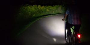 cyclist seen from behind, riding at night on an unlit trail. Only cyclist's rear light and a patch of trail and vegetation illuminated by the cyclist's front light are visible. 