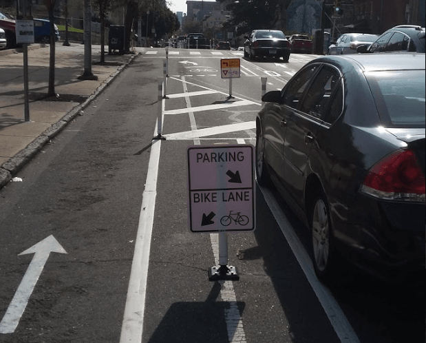 Parking protected bike lane showing parked cars at right, a bike lane against the curb at the left, and between them a sign that says "parking, bike lane" with arrows indicating where to be. 