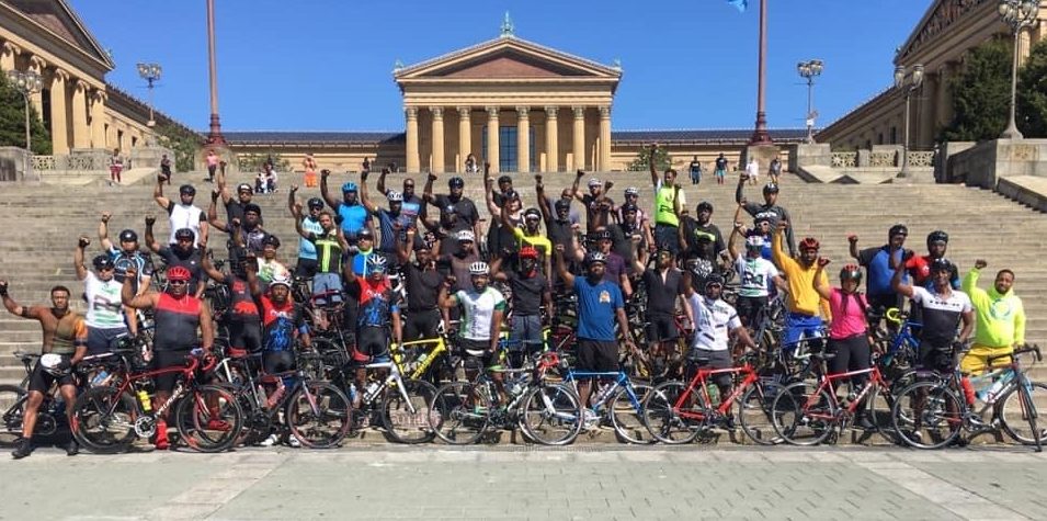 a group of about 100 cyclists pose on the "rocky steps" of the philadelphia museum of art