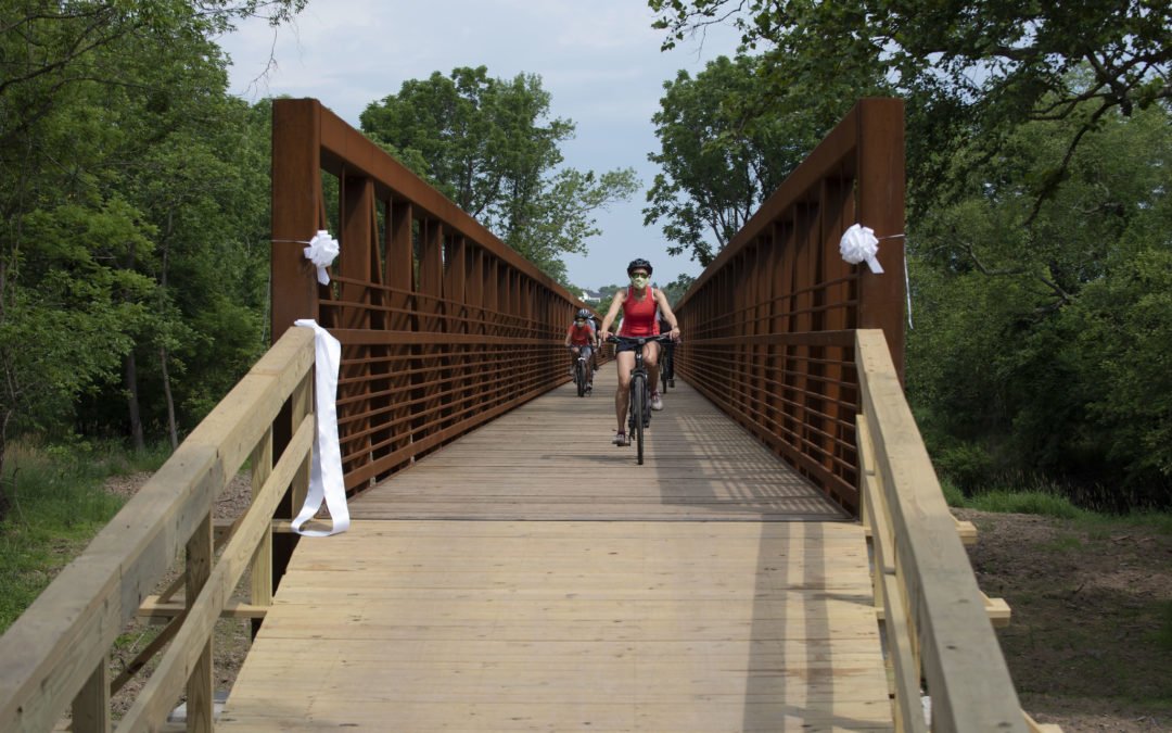 A group of cyclists rides towards the camera on a newly completed steel bridge/boardwalk. The bridge has a temporary wooden ramp connecting it.