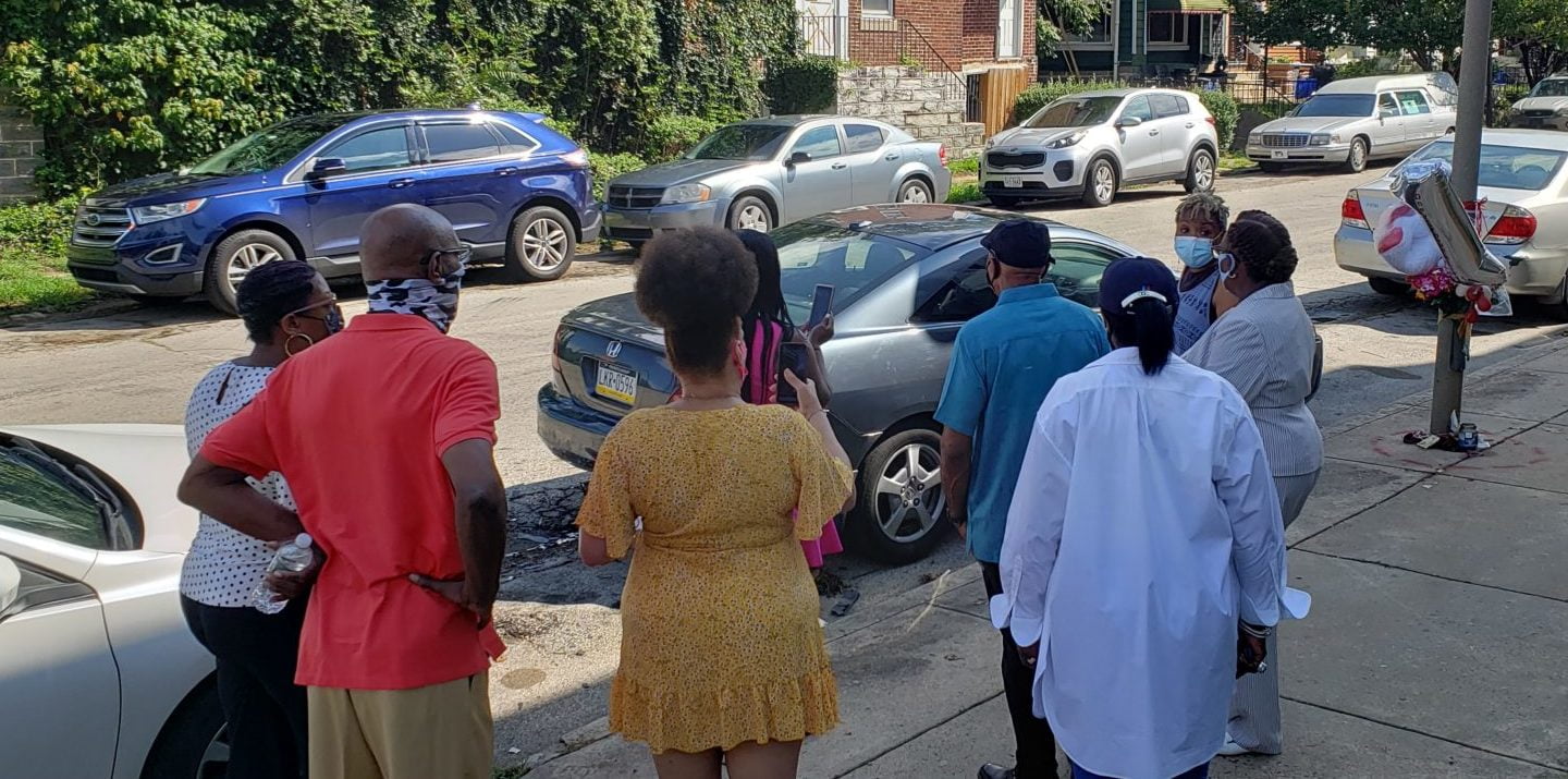 a group of community members stand around the wreckage of a car, with a memorial attached to a light post at righ