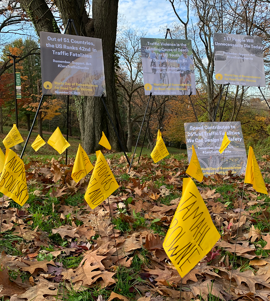 Flags in the ground with names of victims of traffic violence written on them. Signs with traffic violence statistics in the background against a fall sky.