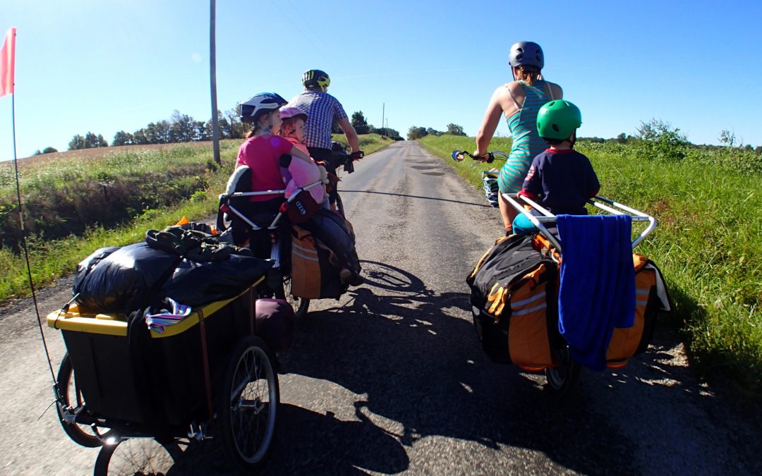 Two adults riding with children in bike trailers on a sunny trail