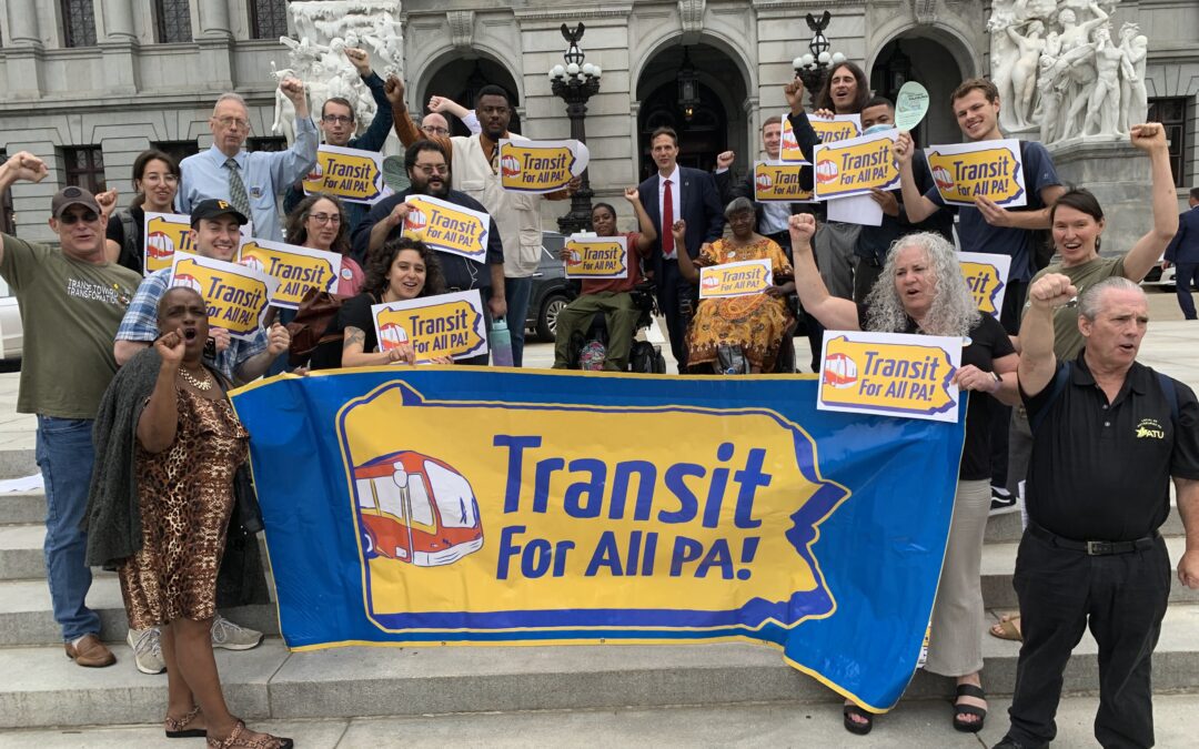 A crowd of 20+ wearing various outfits and of various ethnicities, genders, and abilities with their fists raised, holding yellow and black signs reading "Transit for All PA." They are standing on the steps of the Pennsylvania state capitol
