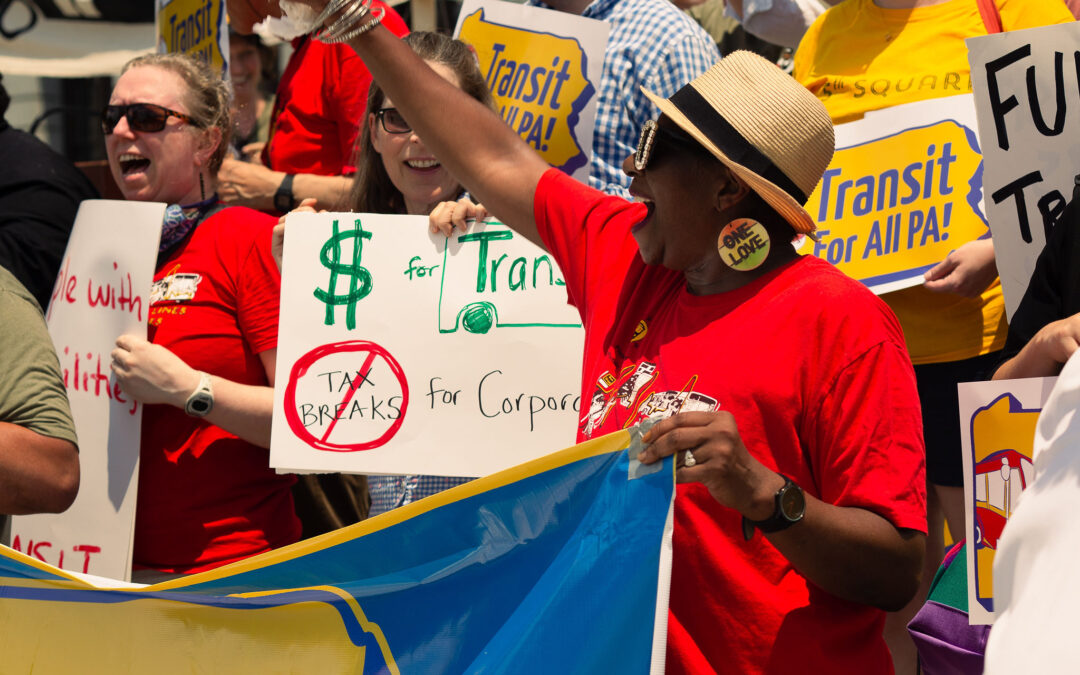 Color image of a Black woman smiling wide and waving her arm while holding a banner. She is wearing a red shirt, rhinestone sunglasses, and a straw hat, in front of a diverse crowd waving yellow signs that read "Transit For All PA"
